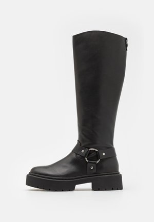 Botte noire en cuir jusqu'aux genoux avec un accent de boucle ronde. Dotée d'une semelle robuste et d'une finition texturée, avec une partie supérieure lisse et une sangle ajustable.