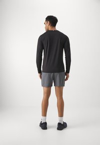 Camisa preta de manga longa para homem com decote redondo, combinada com calções cinzentos. A pessoa está de costas, mostrando um design ajustado e sapatilhas atléticas.