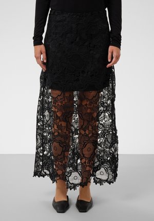 Falda de encaje negra con patrón floral, forro corto por encima de las rodillas, encaje transparente que llega hasta los tobillos, combinada con zapatos planos negros puntiagudos.