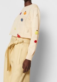 Beżowy sweter dziany z haftowanymi wzorami owoców i liści. Połączony z kremowymi spodniami o dopasowanym kroju i wiązanym pasie.