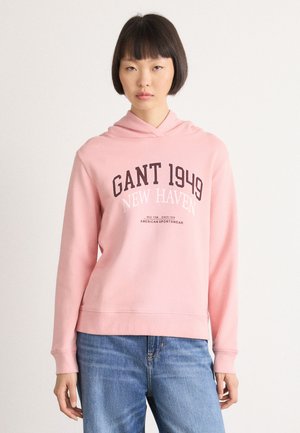 Young woman with short dark hair wearing a pink hoodie with "GANT 1949 New Haven" text and blue jeans, standing against a plain background.