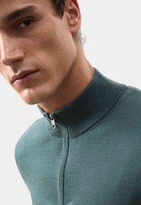 Suéter verde oscuro de cuello alto con una textura suave, que cuenta con un cierre de media cremallera y un cuello acanalado. Vista en primer plano que destaca el ajuste y los detalles.