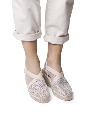 Espadrillas in pizzo di un beige chiaro con ritagli floreali, fascette elastiche incrociate e tacco a zeppa avvolto in juta. Indossate con pantaloni chiari arrotolati.