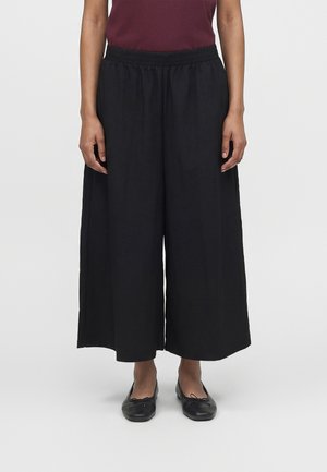 Vrouw draagt losse zwarte wijde broek, bordeauxrood topje en zwarte ballerina's, staand tegen een effen lichte achtergrond.