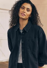 Vrouw met krullend donker haar draagt een zwarte blouse over een grijze top, staand binnen met een neutrale beige achtergrond.