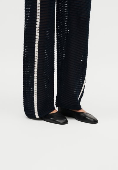 Μαύρα επίπεδα παπούτσια συνδυασμένα με φαρδιά, σκούρα μπλε πλεκτά παντελόνια με λευκές κάθετες ρίγες, που διαθέτουν υφή ανοιχτής πλέξης.