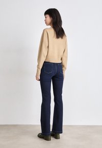 Bež pulover s skrajšanim krojem in rebrastim robom, v kombinaciji s temno denim krilastimi hlačami. Model nosi temno zelene čevlje. Nevtralna ozadje.