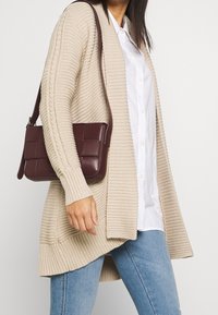 Cardigan beige a coste con maniche lunghe indossato sopra una camicia bianca con bottoni, abbinato a una borsa a tracolla color bordeaux scuro con design trapuntato.
