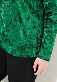 Top in velluto verde con superficie testurizzata, maniche lunghe e vestibilità morbida. Abbinato a jeans neri slim-fit.