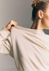 Žena s tetovaním za uchom, ktorá drží golier svojho svetlo krémového rebrovaného svetra, vlasy má zviazané do drdolu, neutrálny pozadie.