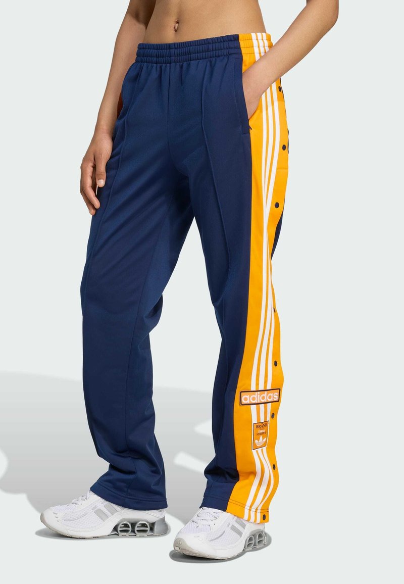 Navy athletic pants with yellow side stripes, snap buttons, and an Adidas logo patch. Made of smooth, breathable fabric.
