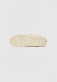 Light beige sneaker outsole with textured grip pattern and circular indentations. Minimal branding on the side. Flat and rounded shape.