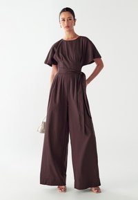 Brun vid byxdress med rund halsringning, korta ärmar, tygbälte och veck vid midjan, tillsammans med en liten ljusfärgad handväska.