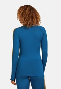 Donna con capelli ricci che indossa un top blu attillato a maniche lunghe e pantaloni coordinati con strisce senape, voltata di spalle su uno sfondo neutro.