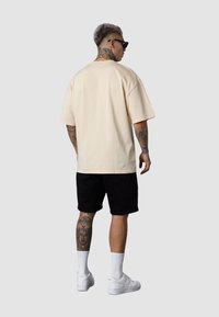 Hellbeiges, oversized T-Shirt mit kurzen Ärmeln, kombiniert mit schwarzen Shorts. Die Person trägt weiße Sneakers und weiße Socken, die komplexe Tattoos zeigen.