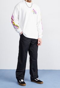 Witte overhemd met lange mouwen en kleurrijke grafische accenten op de mouwen, gecombineerd met zwarte broeken en zwarte sneakers met witte veters en bruine zolen.