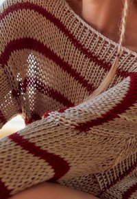 Gros plan d'une personne portant un pull en tricot beige avec des rayures horizontales rouges et un motif à maille ouverte.