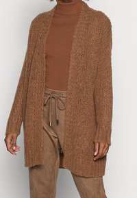 Brun stickad cardigan med öppen framsida, lös passform och ribbade ärmslut. Bärs över en matchande polokrage tröja och mockabyxor.