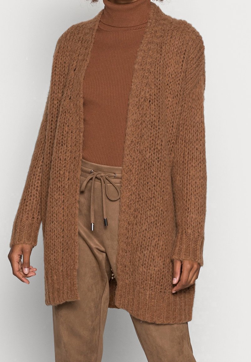 Brun stickad cardigan med öppen framsida, lös passform och ribbade ärmslut. Bärs över en matchande polokrage tröja och mockabyxor.