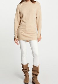 Pull en maille beige avec détails de boutons sur le côté, poignets côtelés et ourlet droit, associé à un jean blanc et des bottes marron décontractées.