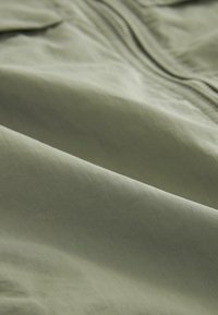 Tissu vert olive clair avec une texture lisse et légèrement réfléchissante, présentant une fermeture éclair visible le long d'un côté. Pas de motifs ou d'accents distincts.