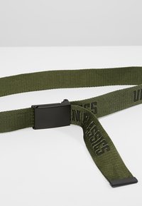 Olīvu zaļa austa josta ar melnu taisnstūra sprādzi, uz kuru ir drukāti burtu „ENDLESS” bolderā fontā gar jostu.