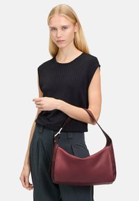 Borsa a mano in pelle bordeaux con forma curva e una corta tracolla, caratterizzata da una texture liscia e una branding discreta.