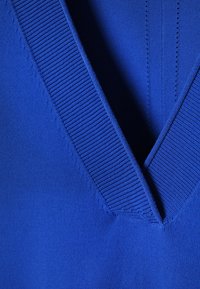 Gros plan sur l'encolure en V d'un pull bleu avec bordure en tricot côtelé et détails de tissu texturé.