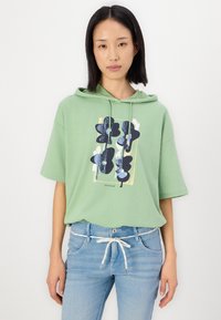 Groene katoenen hoodie met een grafisch bloemenontwerp in zwart en blauw. Korte mouwen, capuchon met trekkoord en een relaxte pasvorm. Gecombineerd met lichte spijkerbroeken.