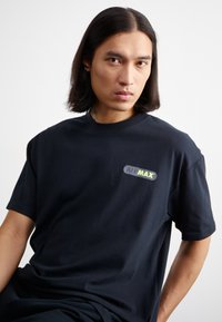Schwarzes Baumwoll-T-Shirt mit kurzen Ärmeln und Rundhalsausschnitt, das ein mehrfarbiges "AIR MAX"-Logo auf der Vorderseite zeigt. Glatte Textur.