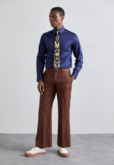Chemise à rayures marines, cravate à motifs, pantalons marron ajustés et baskets blanches avec une semelle beige. Coupe moderne avec des textures nettes.
