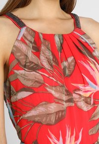 Rød top med tropiske bladmønstre i nuancer af brunt og grønt. Stropper prydet med sorte perler. Let, fladdrende stof.