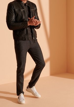Schwarze Lederjacke mit silbernem Zubehör, graue strukturierte Hose und weiße Sneaker. Mann steht mit gefalteten Händen vor einem pfirsichfarbenen Hintergrund.