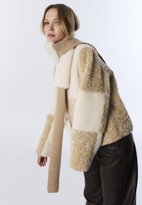 Beige en crème patchwork fleece jas met een geribde gebreide sjaal, met een ontspannen ontwerp en textuuraccenten. Donkerbruine broek daaronder.