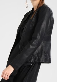 Veste en cuir noir au design ajusté, manches longues et texture lisse. Elle comporte une fermeture éclair à l'avant et une longueur légèrement raccourcie.
