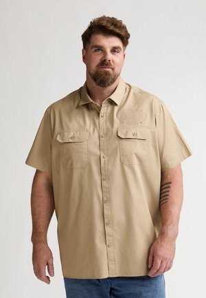 Beżowa koszula z krótkim rękawem, z dwoma kieszeniami na piersi, zapinana na guziki, z teksturowaną naszywką z logo po lewej stronie. Noszona z dżinsami.