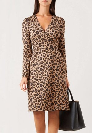 Kobieta w długim rękawie, sukience do kolan w panterkę, trzymająca dużą czarną torbę w prawej ręce.