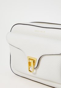 Borsa bianca in pelle con superficie testurizzata, flap curvo, hardware dorato e rifiniture nere. Presenta una chiusura a gancio e branding.