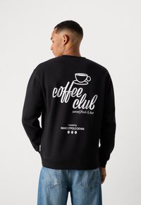 Schwarzes Sweatshirt mit weißem Grafiktext "Kaffeeklatsch" und einer Kaffetasse-Illustration auf dem Rücken. Das Material hat eine glatte Textur.