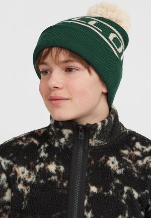 Green knit beanie with white lettering and a fluffy cream pom-pom on top, worn with a zippered black and brown patterned fleece jacket.