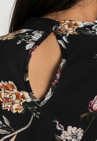Blouse noire avec des motifs floraux en orange, rose et blanc. Le dos présente une découpe en forme de trou de serrure et deux boutons pour la fermeture. Tissu doux et texturé.