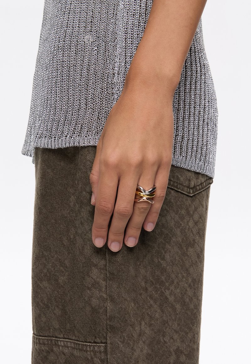Anillo de metal entrelazado en oro, plata y negro en una mano. La mano descansa sobre una tela texturizada de color verde oliva con un patrón sutil.