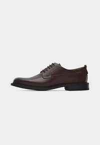 Chaussure habillée pour homme en cuir marron avec semelle noire, lacets à l'avant et languette rouge à l'arrière, vue de profil latéral sur fond blanc.