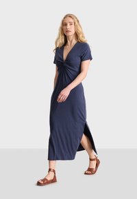 La vestido maxi azul marino, de mangas cortas, con detalle torcido en el frente, abertura lateral, hecho de una tela suave; combinado con sandalias marrones.