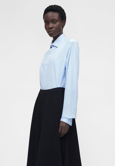 Γυναίκα με κοντά μαλλιά που φοράει ένα ανοιχτό μπλε πουκάμισο με κουμπιά, μπαίνει μέσα σε μια ψηλόμεση μαύρη φούστα, στέκεται μπροστά σε απλό φόντο.