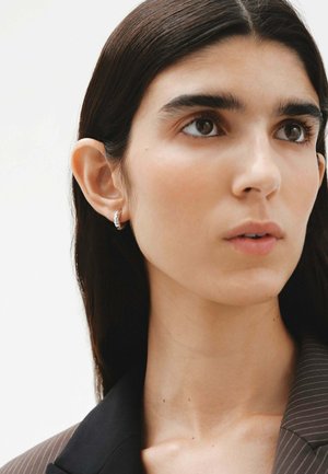 Pendiente de aro de plata con una superficie texturizada, llevado por un modelo con cabello oscuro y liso, vestido con un blazer negro de rayas. Piel clara y maquillaje natural.