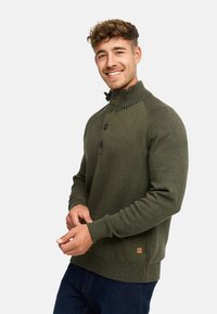 Grøn strikket sweater med høj krave, der har ribbet hals og ærmer, tre-knaps placket og et lille mærke ved kanten.