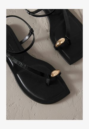 Sandali neri piatti con una finitura lucida, caratterizzati da cinturini sottili e un accento metallico dorato. Design minimalista e texture liscia.