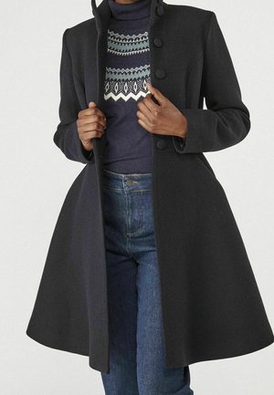 Femme portant un manteau noir mi-long avec de gros boutons, un pull à col roulé bleu foncé à motifs et un jean bleu.