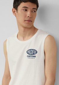 Weißes ärmelloses T-Shirt aus Baumwolle mit einem blauen ovalen Logo und dem Text "PACIFIC CUSTOMS BOARDS&KITS" auf der Vorderseite.